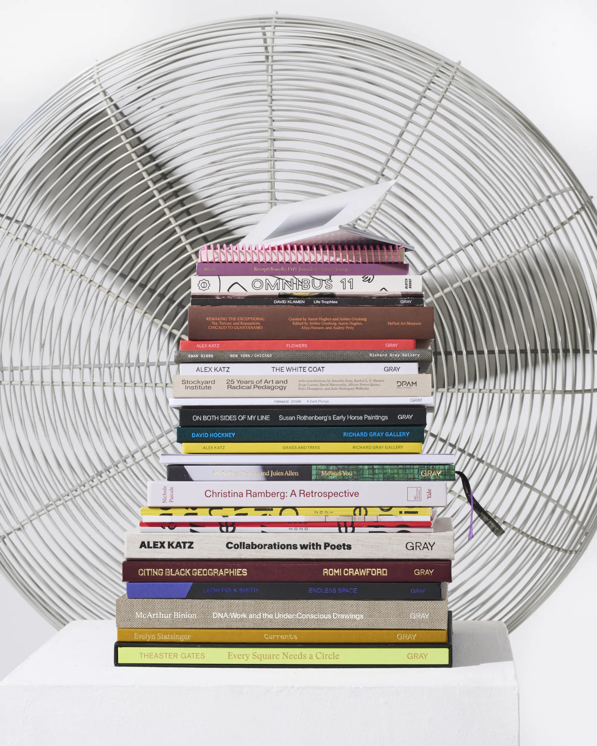 An image of several Hour Studio designed books stacked on top of each other, on top of an office chair, in front of a large fan.